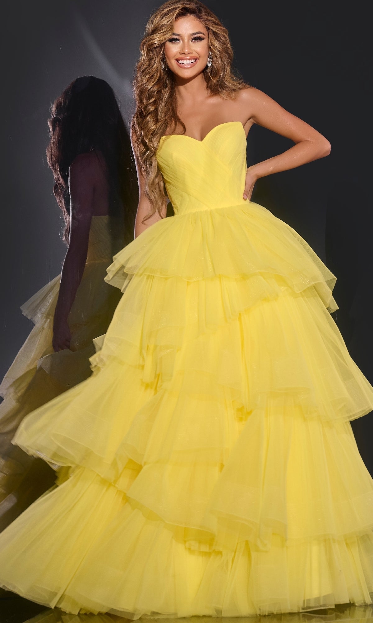 A woman smiles in the Jovani 40394 Strapless Ruffled Prom Ball Gown, a bright yellow tiered tulle dress, posing against a dark background with her reflection beside her.